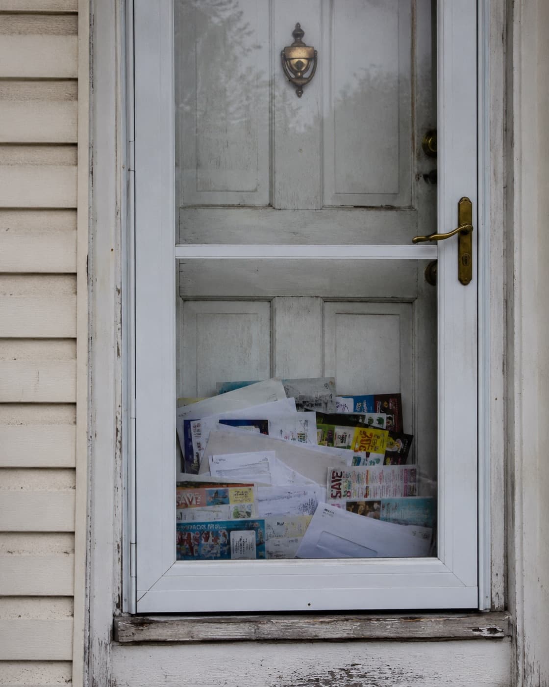 A closed front door behind a storm door, with mail and delivery flyers piled up against the glass where nobody has collected them.