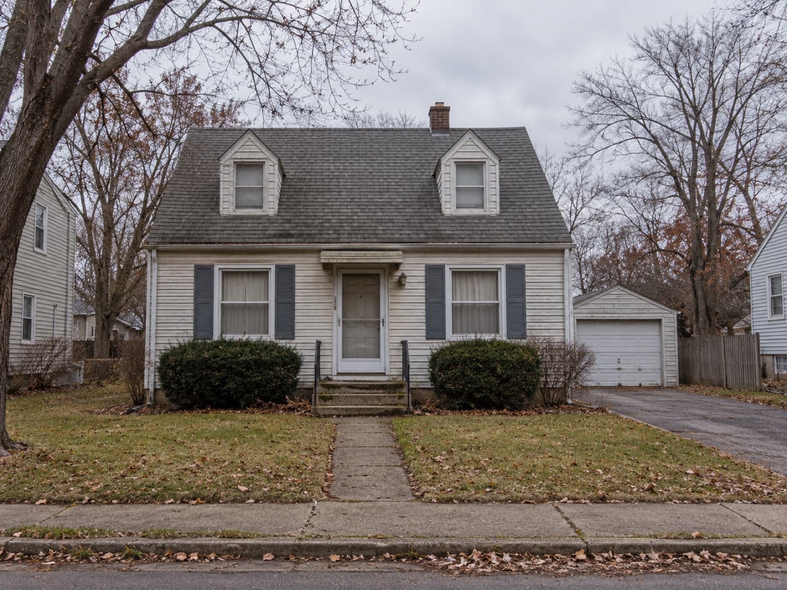 A post war Cape Cod house on a residential street, photographed straight on from the sidewalk on an overcast day. The lawn is covered in fallen leaves, the curtains are drawn and there are no cars on the driveway.